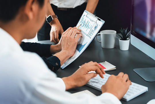 techstrategy-img-blurple Two coworkers analyzing data, with one pointing to charts on a clipboard while another works at a keyboard in an office setting.